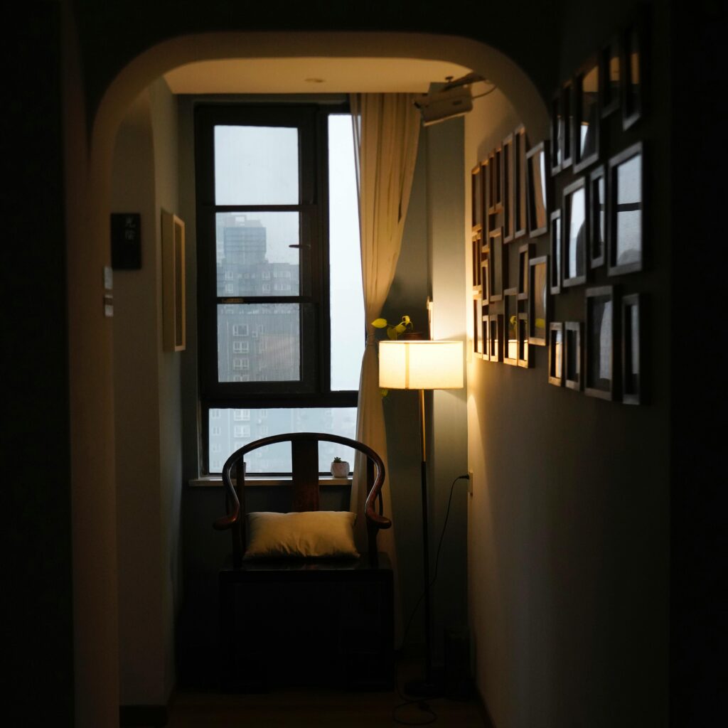 A dimly lit cozy corner in an apartment with a chair, lamp, and window view.